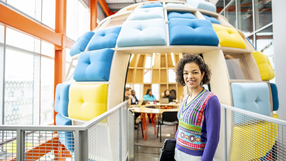 A student stood outside of the Here East pod and a group of students sat at a table in the background.