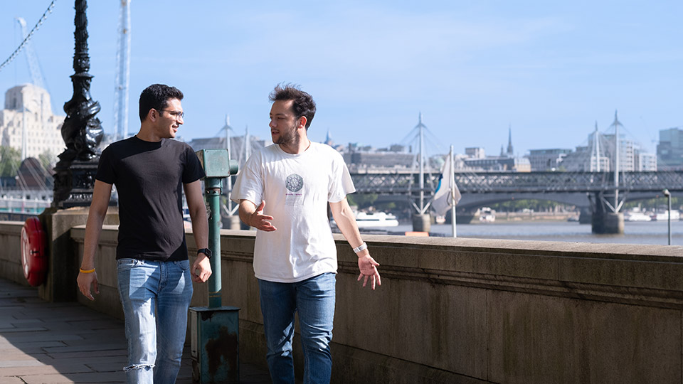 Two students walking along London South Bank with the river and buildings in the background.