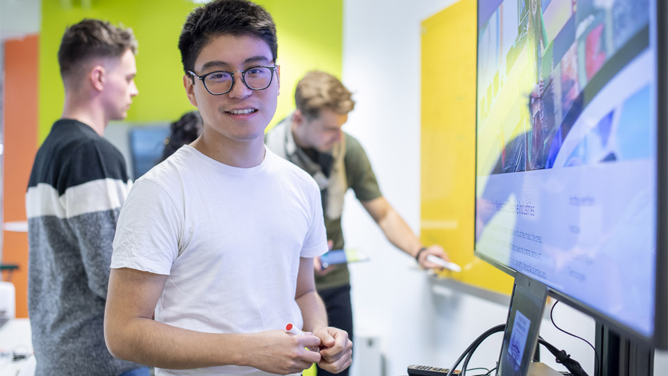 A student stood in front of a computer screen with people working in the background.