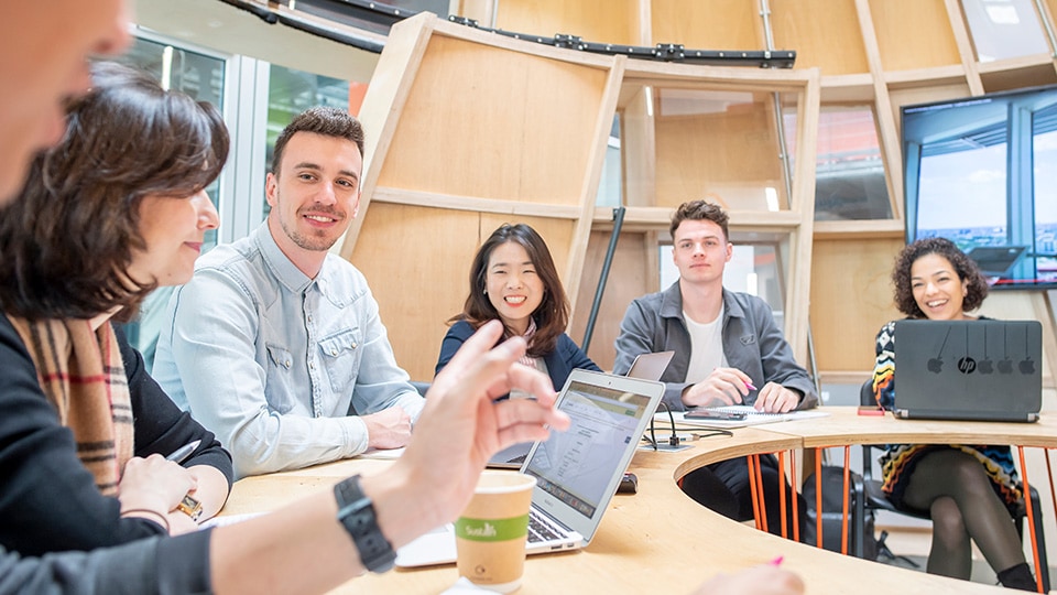 Six people sitting at a round table talking and using their laptops