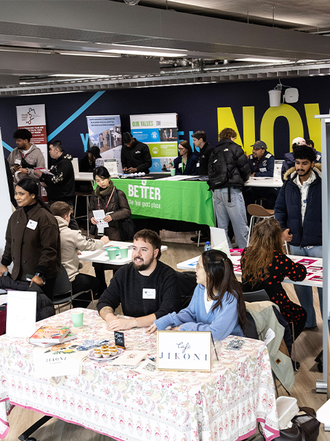 A busy Student Job Fair at Here East, where people are gathered around tables, chatting and engaging with exhibitors.