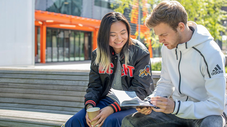 Two students sat on a bench and one holding a notebook.