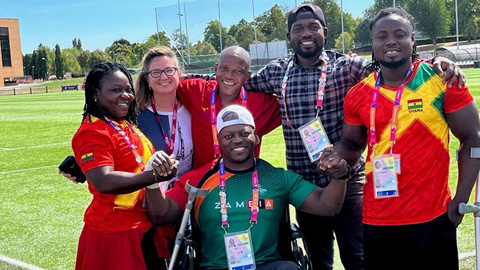 6 people outside, in front of a Rugby pitch, including Para athletes and coaches from Ghana and Zambia a practitioner from Loughborough smiling for the camera